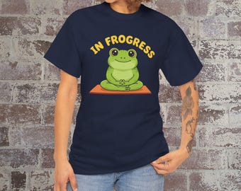 May include: Navy blue t-shirt featuring a cartoon frog meditating on an orange mat. The text "IN FROGRESS" is arched above the frog in yellow. The shirt is a short-sleeved crew neck.