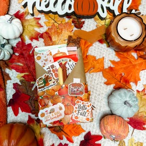 Puede incluir: Un arreglo con temática otoñal con calabazas decorativas, hojas de otoño y una vela. Las palabras "hello fall" se muestran en un recorte de madera. Un sobre de papel kraft está decorado con pegatinas que dicen "Just a girl who loves fall" y "Hello Fall".