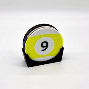 May include: A set of round coasters with a billiards theme. Each coaster has a yellow and white design with the number 9 in black. The coasters are held in a black, angled holder.