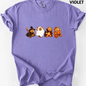 May include: A violet short-sleeved t-shirt featuring a graphic of four teddy bears dressed in Halloween costumes: a witch with a broom, a ghost, a vampire with a lantern, and a pumpkin. The text VIOLET is printed in the top right corner.