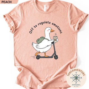 May include: Heather peach-colored t-shirt featuring a cartoon goose riding a scooter and holding a coffee cup. The text "Off to regulate emotions" is arched above the goose. The shirt has a crew neck and short sleeves. The Threadventure Apparel logo is in the bottom right corner.