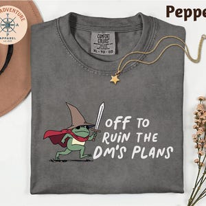 May include: Pepper-colored t-shirt with a graphic of a frog wearing a wizard hat and holding a sword. Text reads "off to ruin the DM's plans." A gold star pendant necklace is draped over the shirt.
