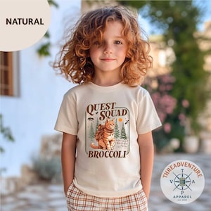 May include: A beige t-shirt featuring a graphic of a cat with a backpack, the words "QUEST SQUAD BROCCOLI", and the text "So Cute, Isn't It, And It's Good For You". The shirt is worn by a child.