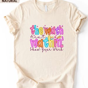 May include: A natural-colored t-shirt featuring a colorful graphic print with the text "the math Ain't mathin' Show Your Work". The design includes bright pink, yellow, orange, and blue bubble letters, small flower icons, a stack of books, and a pencil.