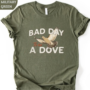 May include: A military green short-sleeve t-shirt featuring a graphic of a flying dove in the center. The text "BAD DAY" appears above the bird, "To be" is written in a smaller red font, and "A DOVE" is printed below the bird in white.