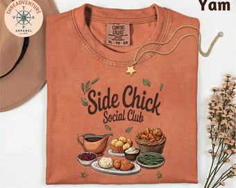 May include: A burnt orange t-shirt with the text "Side Chick Social Club" and a graphic of Thanksgiving food. A gold star pendant necklace is draped over the shirt. The shirt has a Comfort Colors tag.