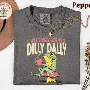 a gray t-shirt with a frog sitting on a mushroom and holding a cup of coffee, accompanied by a necklace and a hat.