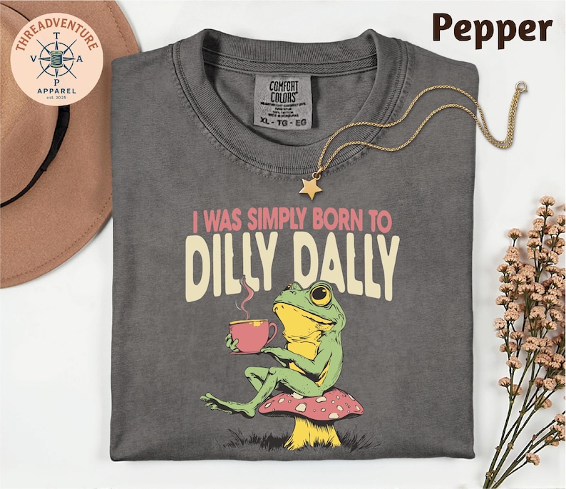 a gray t-shirt with a frog sitting on a mushroom and holding a cup of coffee, accompanied by a necklace and a hat.