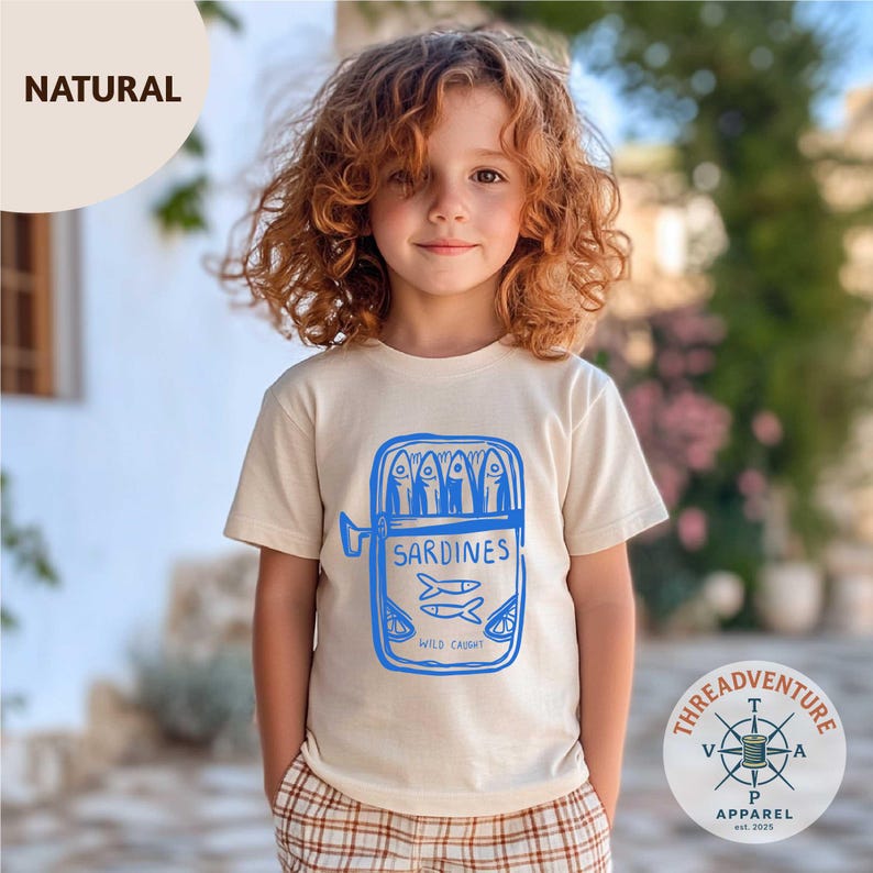 May include: A beige t-shirt with a blue graphic of a sardine can, featuring the word "SARDINES" and "WILD CAUGHT". The shirt is worn by a child with curly red hair. The shirt has a round neck and short sleeves.