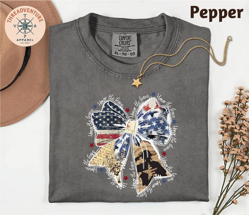 a gray t-shirt with a patriotic design, including a bow and stars, along with a necklace and a hat.
