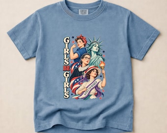 May include: A blue jean colored short-sleeve t-shirt featuring a graphic print of four figures including the Statue of Liberty and a woman holding a torch with a sash reading VOTES FOR WOMEN. The text GIRLS BE GIRLS is printed vertically in black.