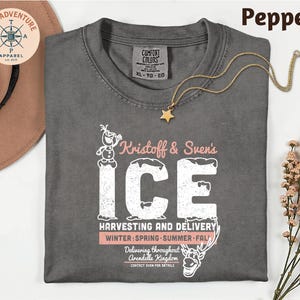 A gray t-shirt with a graphic design featuring a character from the Disney movie Frozen, along with text that says "Knostoff & Sven's ICE". The t-shirt is laid out on a white surface, with a hat and some flowers nearby.
