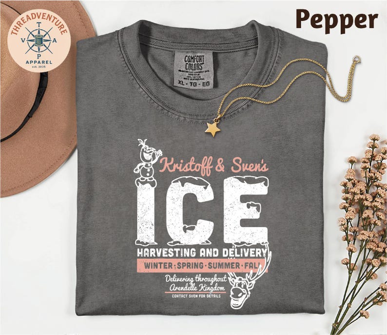 A gray t-shirt with a graphic design featuring a character from the Disney movie Frozen, along with text that says "Knostoff & Sven's ICE". The t-shirt is laid out on a white surface, with a hat and some flowers nearby.