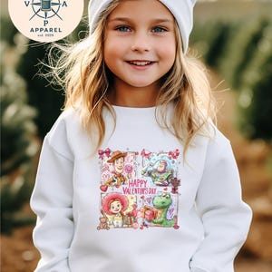 A young girl wearing a white sweater with a colorful graphic of characters from Toy Story and Happy Valentine's Day.