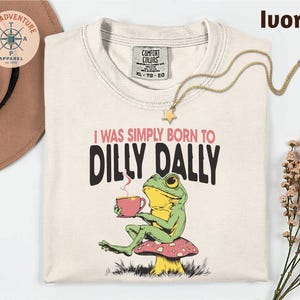 a white t-shirt with a colorful frog graphic and text, along with a hat, a necklace, and some flowers.