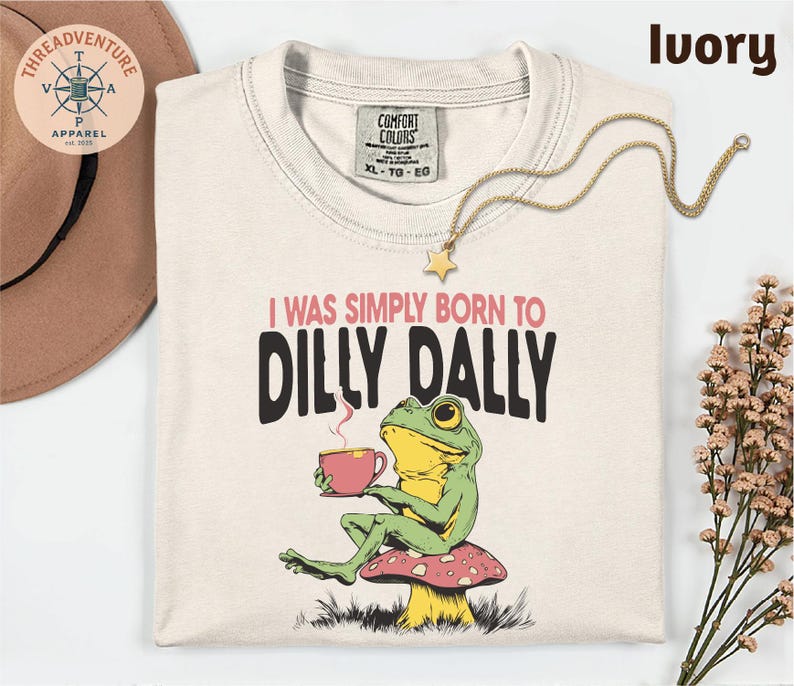 a white t-shirt with a colorful frog graphic and text, along with a hat, a necklace, and some flowers.