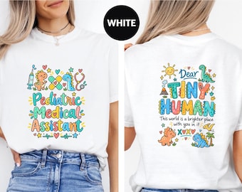 May include: A white t-shirt featuring a colorful graphic print on the front and back. The front displays the text Pediatric Medical Assistant with dinosaur and medical icons. The back features the text Dear Tiny Human This world is a brighter place with you in it alongside various dinosaur illustrations and a sun.