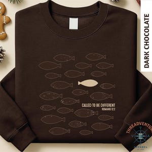 A brown long-sleeved t-shirt with a graphic design of fish and a single fish in the center, along with text that reads "CALLED TO BE DIFFERENT ROMANS 12:2".