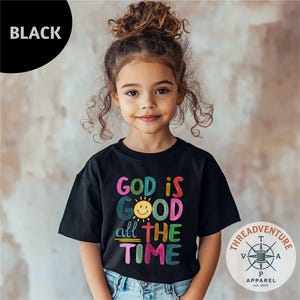 May include: A black t-shirt with the text "GOD IS GOOD all THE TIME" in colorful, playful lettering. The design includes a smiling sun graphic. The shirt is from Threadventure Apparel, est. 2025.