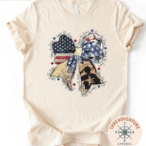 a white t-shirt with a graphic design depicting a large bow made up of various elements, including stars, stripes, and a flag. The bow is surrounded by text that appears to be a patriotic theme, possibly referencing American symbols and values.
