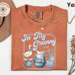 a red t-shirt with a graphic design of a duck and various kitchen items, including a teapot, bowls, and a star, along with the text "In My Granny era".
