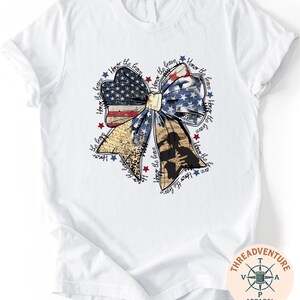 A white t-shirt with a graphic design featuring a large bow made up of various elements, including stars, stripes, and a flag. The bow is surrounded by text that appears to be a patriotic theme or a tribute to the United States.