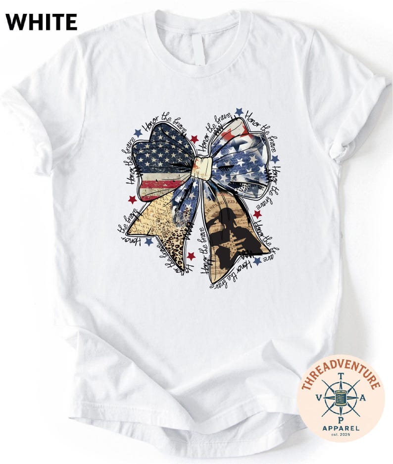 A white t-shirt with a graphic design featuring a large bow made up of various elements, including stars, stripes, and a flag. The bow is surrounded by text that appears to be a patriotic theme or a tribute to the United States.
