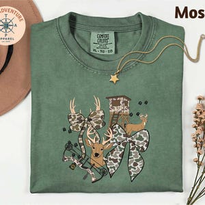 May include: A moss green Comfort Colors t-shirt with a hunting-themed graphic. The graphic includes a deer, bow, camouflage bows, and a hunting blind. A gold star pendant necklace is draped across the shirt.