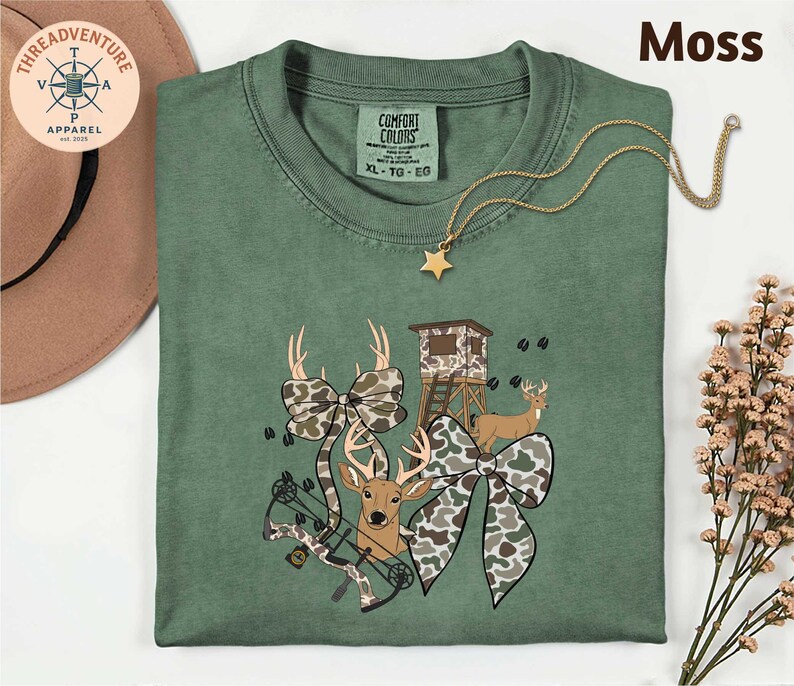 May include: A moss green Comfort Colors t-shirt with a hunting-themed graphic. The graphic includes a deer, bow, camouflage bows, and a hunting blind. A gold star pendant necklace is draped across the shirt.