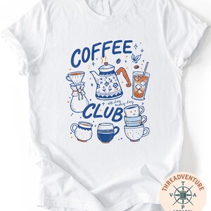 A white t-shirt with a graphic design featuring various coffee-related items such as a coffee pot, coffee cup, and iced coffee cup, along with the text "COFFEE CLUB" and other related illustrations.