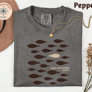 a gray t-shirt with a graphic design of fish, one of which is different from the rest, and the text "Called to be different" along with a Bible verse reference. The t-shirt is laid out on a white surface, accompanied by a hat, a necklace, and some flowers.