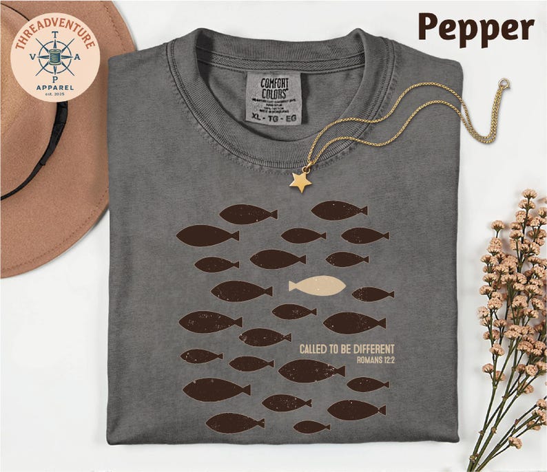 a gray t-shirt with a graphic design of fish, one of which is different from the rest, and the text "Called to be different" along with a Bible verse reference. The t-shirt is laid out on a white surface, accompanied by a hat, a necklace, and some flowers.