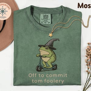 May include: Moss green t-shirt with a graphic of a frog wearing a wizard hat and riding a scooter. The text "Off to commit tom foolery" is printed below the graphic. A gold star pendant necklace is draped on the shirt.