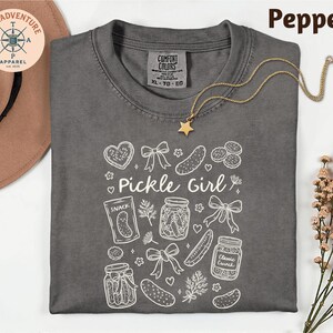 a gray t-shirt with a white design, including various pickle-related items such as jars, a star, and a heart. The t-shirt is laid out on a white surface, with a hat and some flowers nearby.