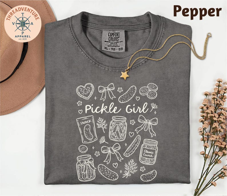 a gray t-shirt with a white design, including various pickle-related items such as jars, a star, and a heart. The t-shirt is laid out on a white surface, with a hat and some flowers nearby.