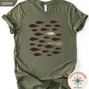A military green t-shirt with a graphic design of a group of fish and a single fish in the center, along with the text "Called to be different" and "Bible verse 12:22".