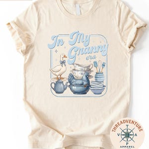 A t-shirt with a graphic design featuring a white duck, a blue teapot, and various kitchen utensils such as spoons and bowls.