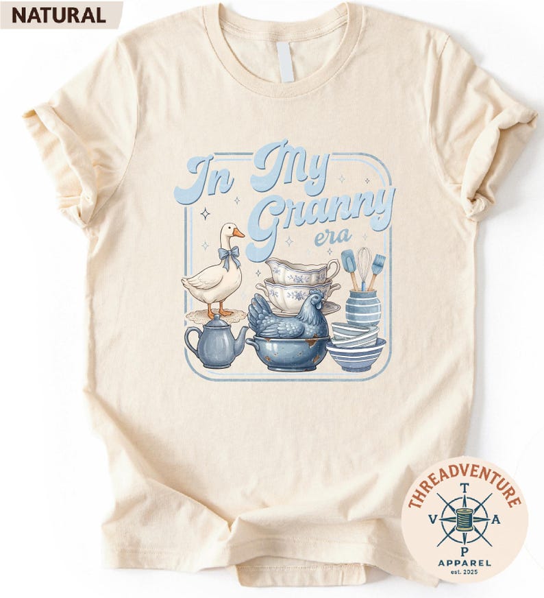 A t-shirt with a graphic design featuring a white duck, a blue teapot, and various kitchen utensils such as spoons and bowls.