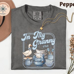 a gray t-shirt with a graphic design depicting a duck surrounded by various kitchen items, including a teapot, a bowl, and a spoon. The text on the t-shirt reads "In My Granny era".