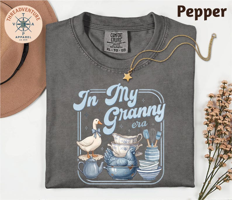 a gray t-shirt with a graphic design depicting a duck surrounded by various kitchen items, including a teapot, a bowl, and a spoon. The text on the t-shirt reads "In My Granny era".