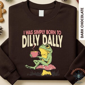 A brown long-sleeved t-shirt with a graphic design depicting a green frog sitting on a mushroom and holding a cup of coffee.