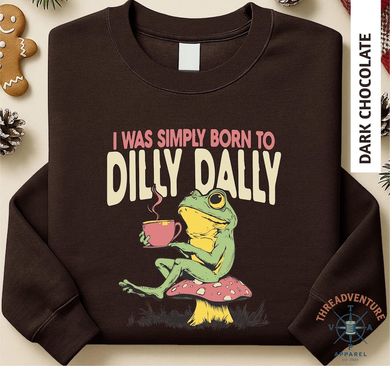 A brown long-sleeved t-shirt with a graphic design depicting a green frog sitting on a mushroom and holding a cup of coffee.