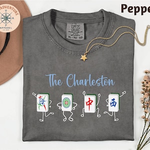 a gray t-shirt with a design of four Chinese tiles arranged in a row, each depicting a different character. The t-shirt is laid out on a white surface, with a hat and some flowers nearby.