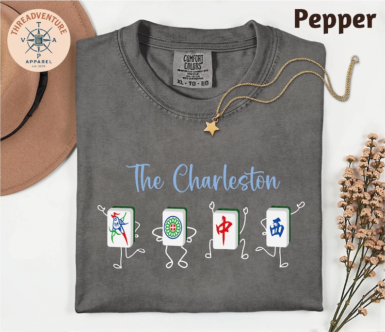 a gray t-shirt with a design of four Chinese tiles arranged in a row, each depicting a different character. The t-shirt is laid out on a white surface, with a hat and some flowers nearby.