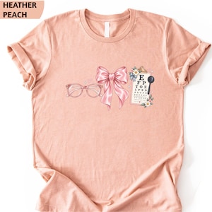 May include: A heather peach t-shirt featuring a graphic print of pink glasses, a large pink bow, and an eye chart with the letters E, F, P, T, O, Z, L, P, E, D, E, D, F, C, E, P, F, E, L, O, P, D, E, D, F, E, F, C, E, P, and a black occluder.
