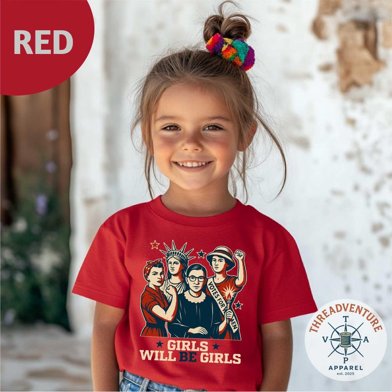 May include: A young girl wearing a red t-shirt with a graphic of women and the text "GIRLS WILL BE GIRLS". The graphic includes the Statue of Liberty and the text "VOTES FOR WOMEN". The shirt has a Threadventure Apparel logo.