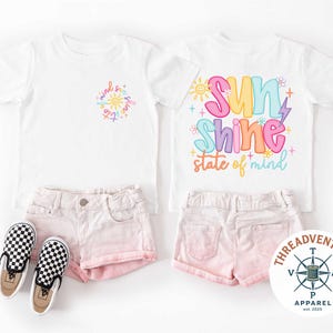 以下が含まれることがあります： カラフルなグラフィックとピンクのショートパンツが特徴の白いTシャツ2枚。1枚のシャツには「sunshine state of mind」、もう1枚には「sunshine」と書かれています。白と黒のチェック柄のスリッポンシューズも画像にあります。