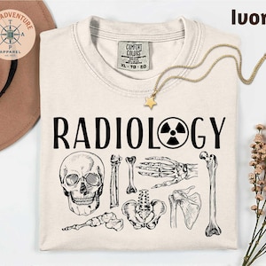 May include: Ivory-colored t-shirt with the word "RADIOLOGY" in bold black letters, accompanied by a radiation symbol and detailed illustrations of human skeletal elements. A gold necklace with a star pendant is draped across the shirt.