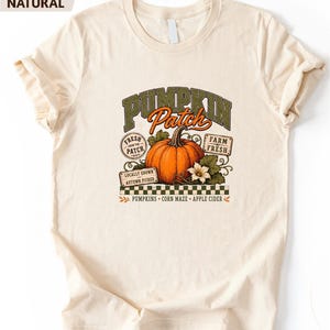 May include: A natural-colored t-shirt featuring a graphic of an orange pumpkin with green leaves and white flowers. The text reads PUMPKIN Patch, FRESH FROM THE PATCH, FARM FRESH, LOCALLY GROWN AUTUMN PICKED, PUMPKINS, CORN MAZE, and APPLE CIDER.