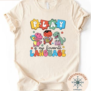 May include: A cream-colored t-shirt with a colorful graphic featuring the word "PLAY" in puzzle piece letters above three cartoon dinosaurs. The phrase "is my favorite LANGUAGE" is below the dinosaurs. A small logo is in the bottom right corner.